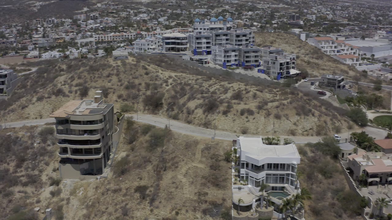 Elevated Los Cabos view with a round multi-story house in the foreground, a pale roofed home below it, and white apartment buildings across the ridge, with no visible people.