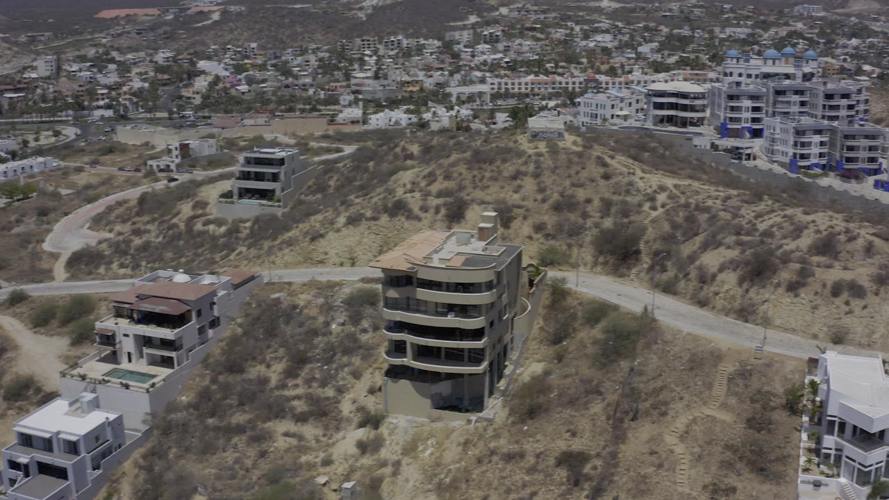 Aerial frame over Los Cabos, Mexico, showing dry hillside terrain, a curved dirt road, modern homes in the foreground, distant apartment buildings, and no visible people.