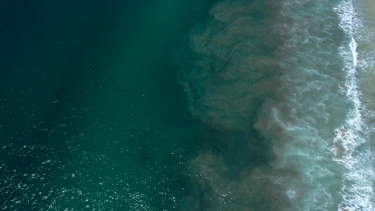 Aerial frame over Los Cabos water showing white foam crossing a pale shallow patch beneath the green surface, with no visible people.