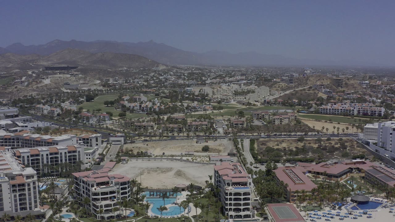Opening aerial frame over Los Cabos, Mexico, with resort buildings and blue pools in the foreground, city development beyond, hazy mountains on the horizon, and no visible people.