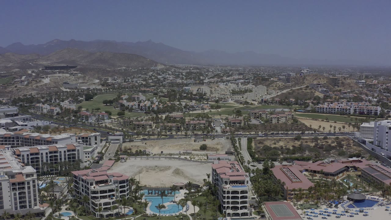 Los Cabos aerial view one second in, showing hotel roofs and pools near the bottom edge, a broad road corridor through the middle, low buildings beyond, and no visible people.