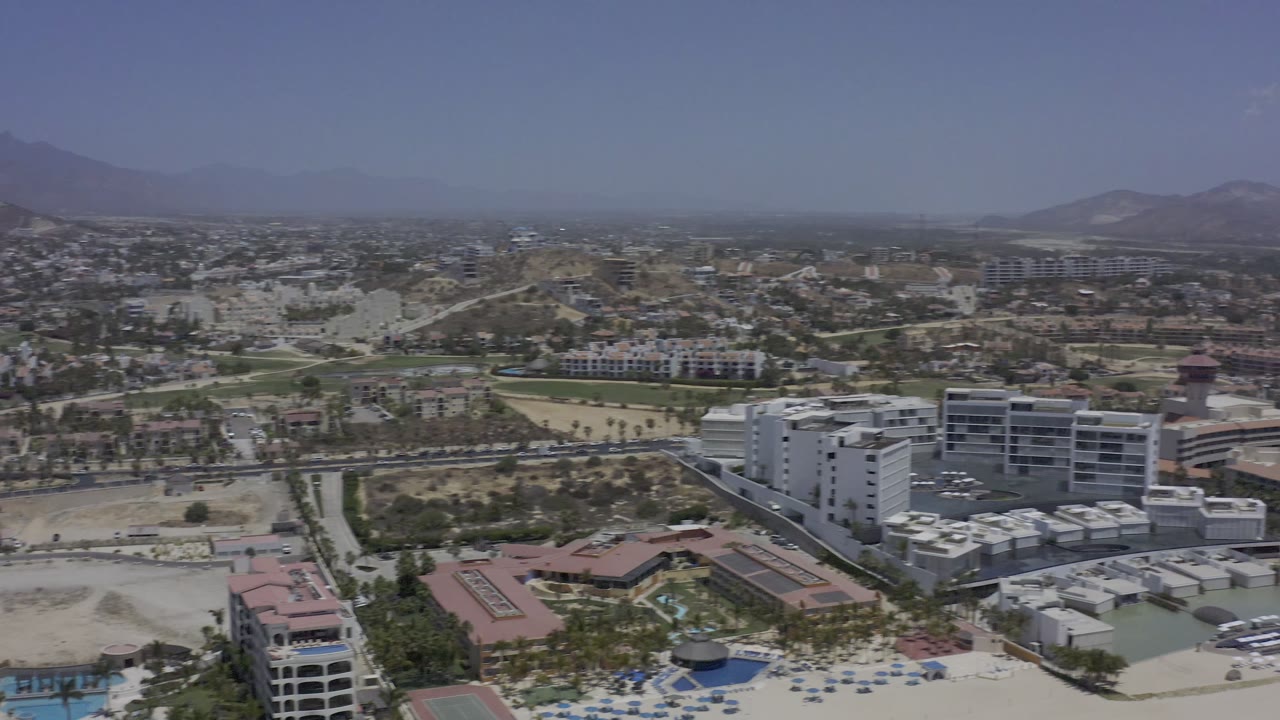 Wide Los Cabos aerial frame with white multi-story buildings in the foreground, roads through a dry valley, distant neighborhoods, mountains, and no visible people.