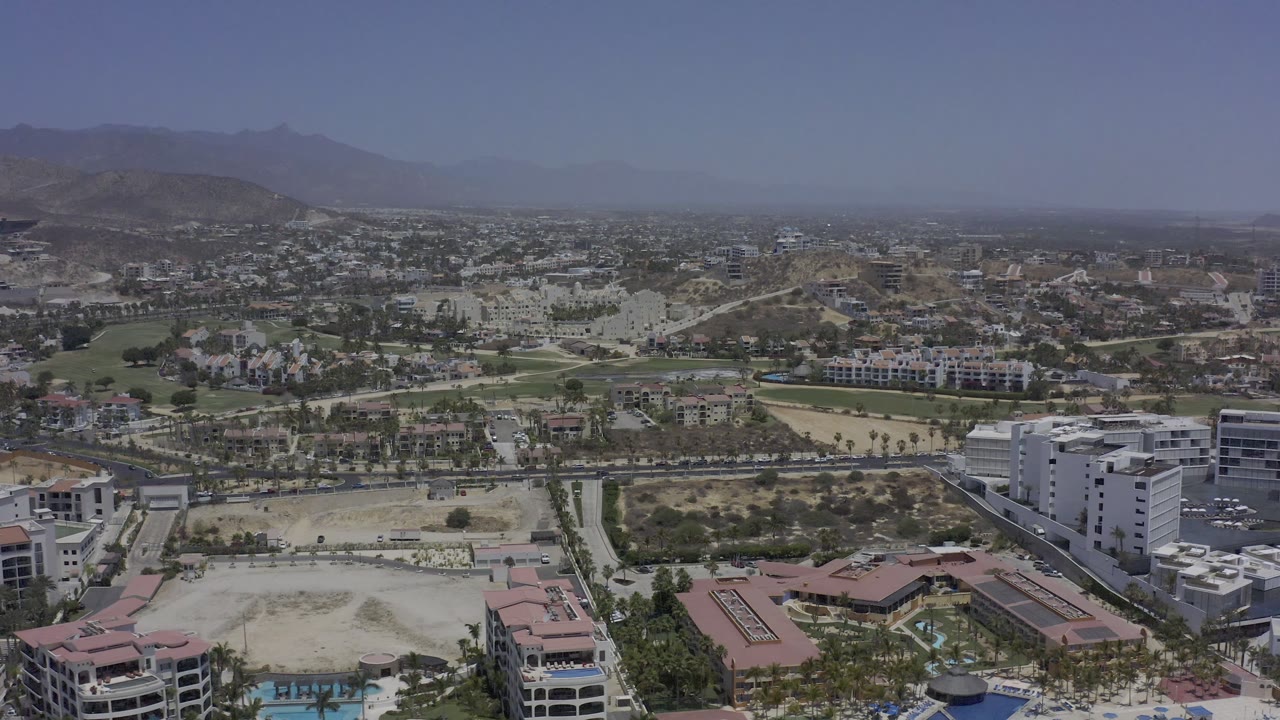 Los Cabos aerial view with white multi-story buildings in the foreground, landscaped fairways and roads in the middle distance, hazy mountains, and no visible people.