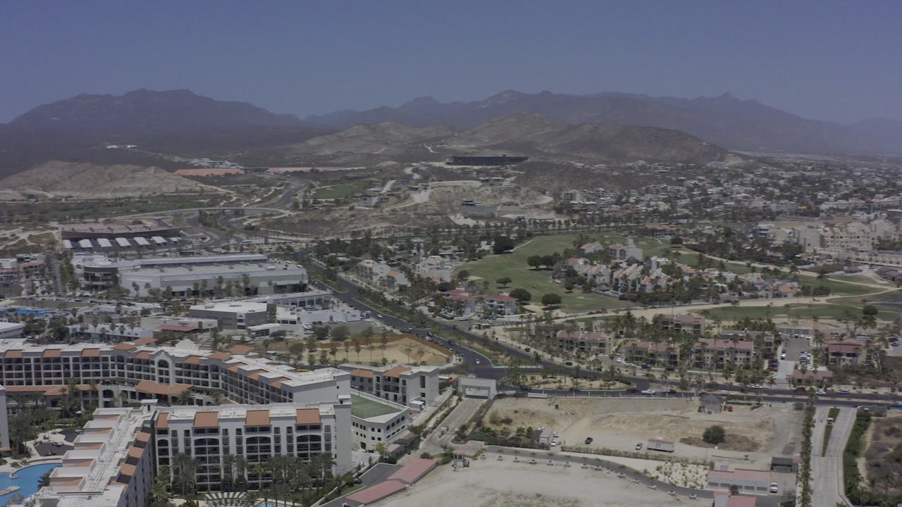 Wide aerial frame over Los Cabos with resort buildings in the foreground, curving roads and green grounds in the middle distance, hazy mountains beyond, and no visible people.