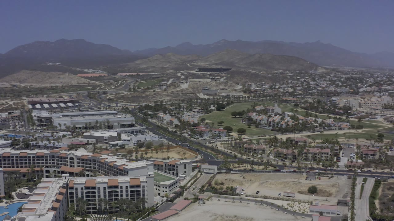 Los Cabos aerial view with hotel-style buildings at the bottom edge, a road corridor crossing the frame, green resort grounds, dry terrain, distant mountains, and no visible people.