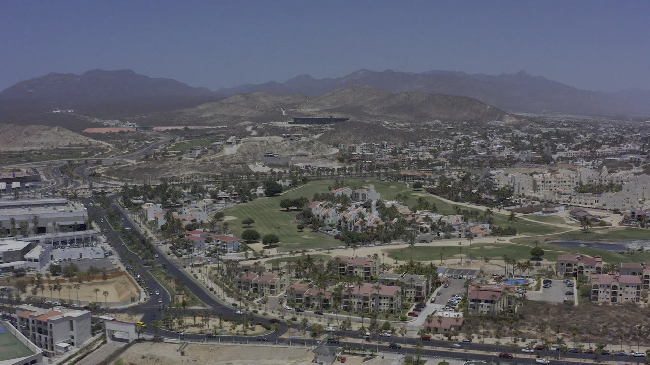 Wide aerial view over Los Cabos with paved roads, low-rise buildings, green fairways, dry terrain, distant mountains, and no visible people.