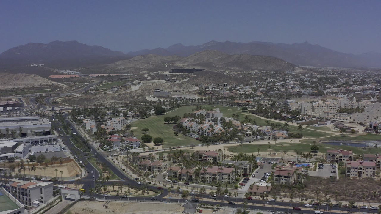 Aerial frame in Los Cabos showing a main road, scattered cars, resort-style buildings, landscaped fairways, desert lots, mountain ridges, and no visible people.