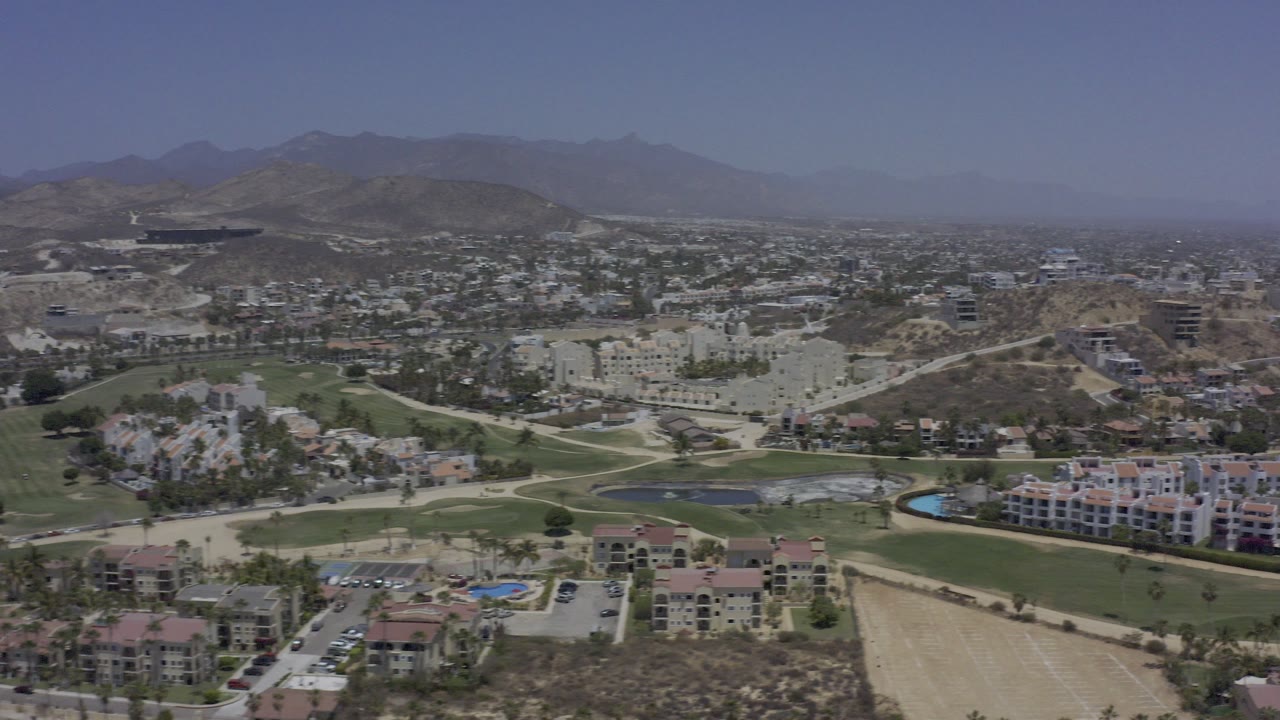Aerial view over Los Cabos buildings, golf greens, roads, dry hills, and hazy mountains.