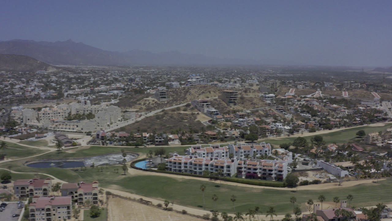Opening aerial frame over Los Cabos with green fairways in the foreground, low buildings and roads across the midground, hazy mountains in the distance, and no visible people.