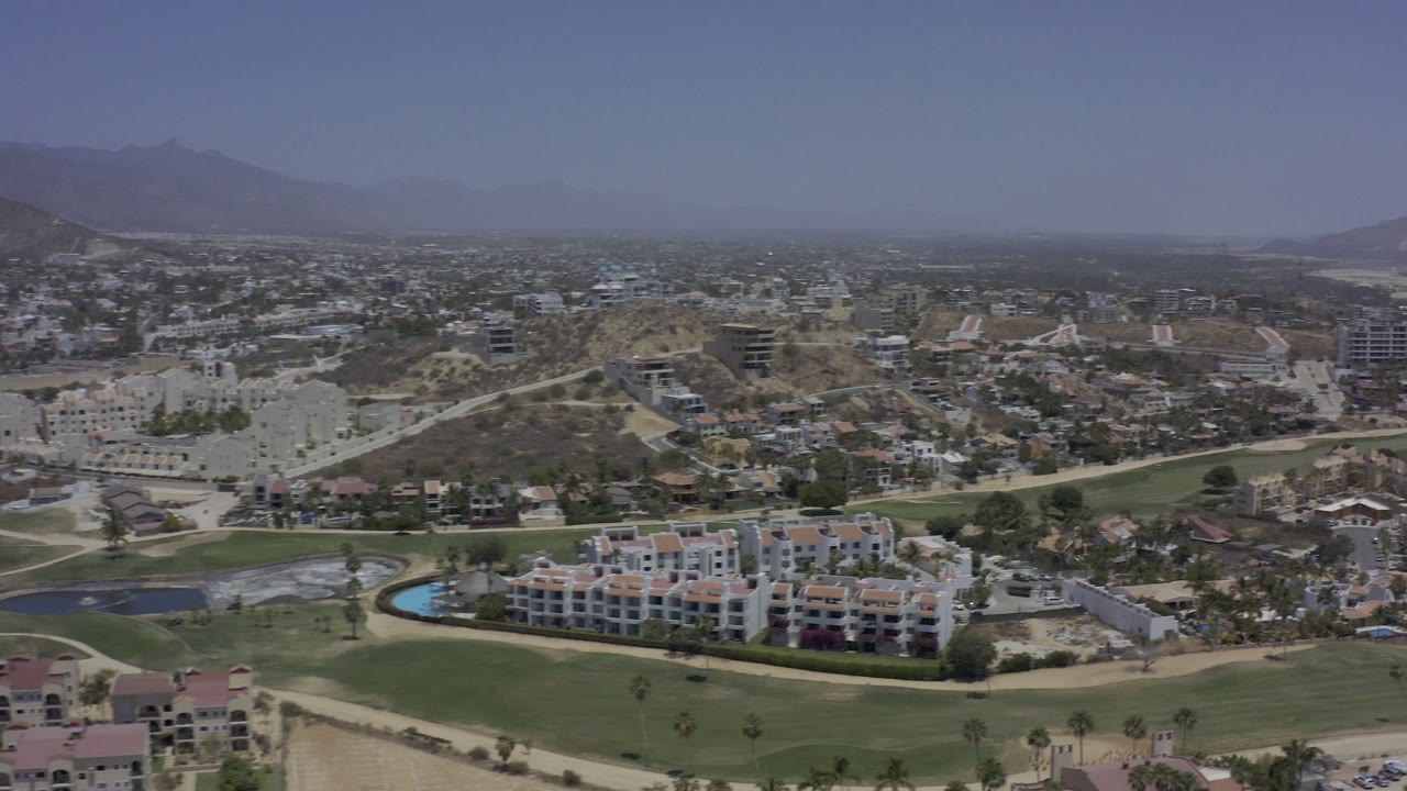 High aerial view of resort-style buildings, green course land, roads, and mountain backdrop.