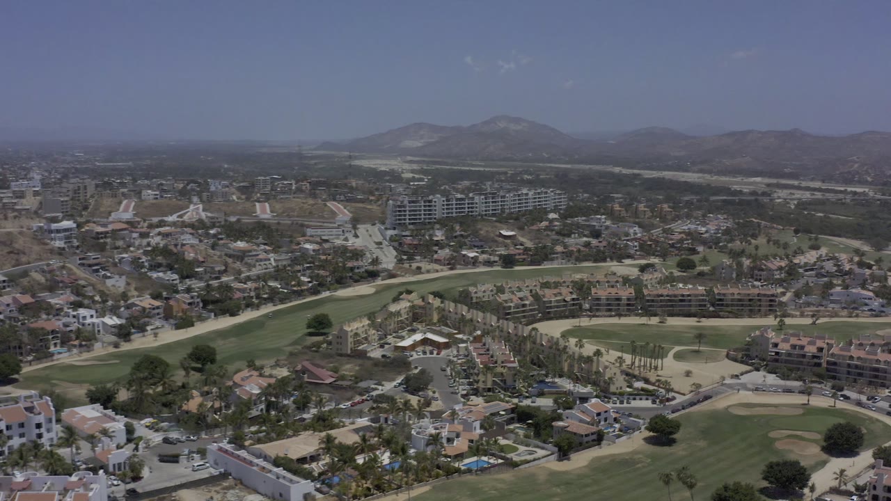 Aerial frame in Los Cabos, Mexico, looking across clustered homes, a broad green fairway, larger buildings near the center, hazy hills, and no visible people.