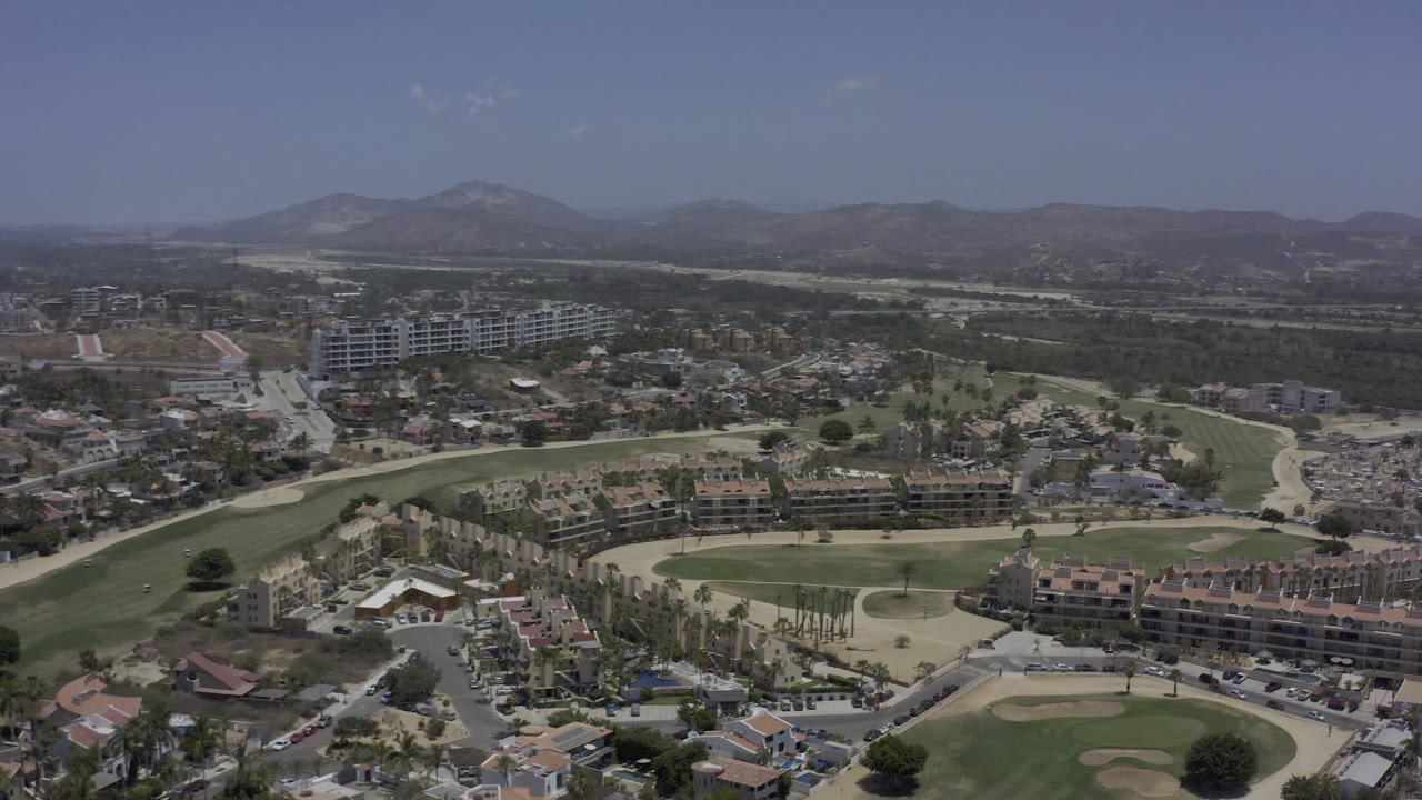 The second Los Cabos aerial frame keeps the golf course in the lower frame, with apartment-style buildings, roads, palms, and a hazy mountain ridge under a clear blue sky, with no visible people.