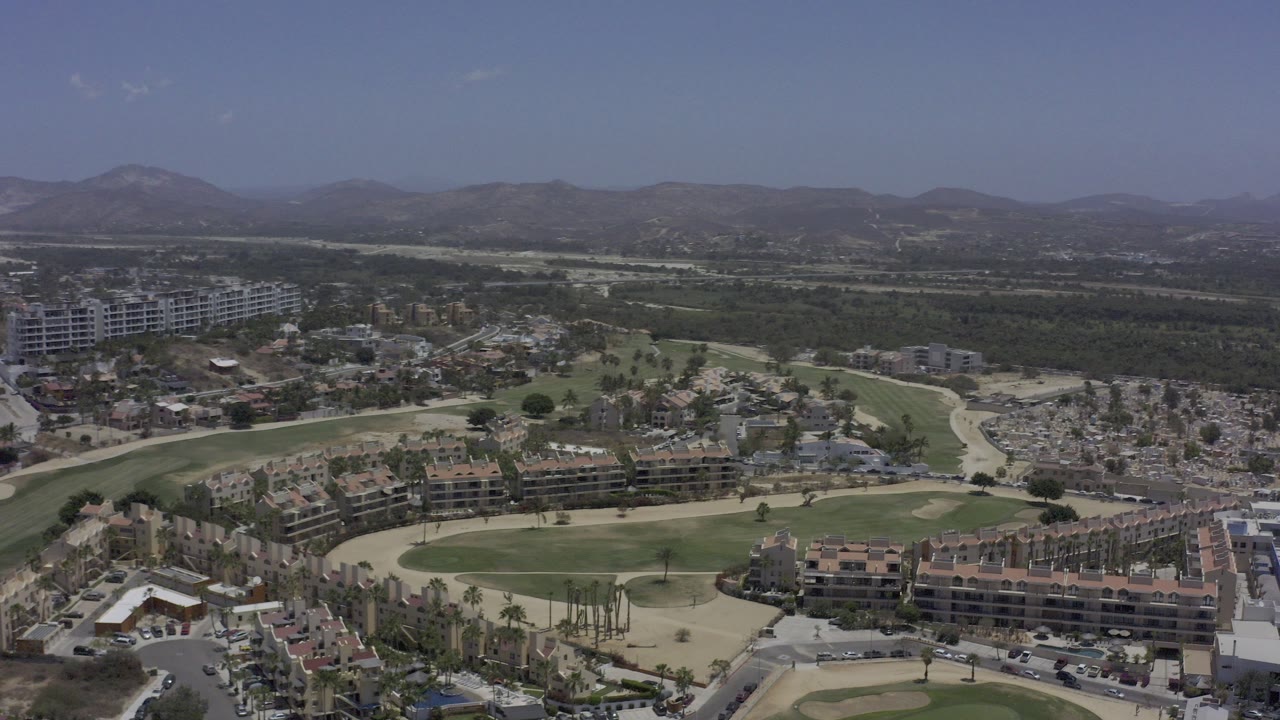 High aerial view in Los Cabos, Mexico, looking over apartment complexes beside green lawns, roads, dry terrain, and distant low mountains, with no visible people.