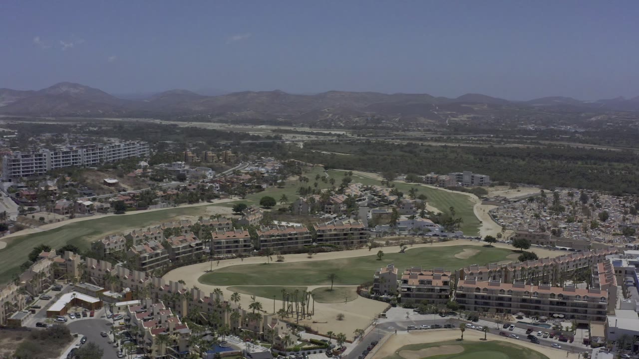 Aerial frame in Los Cabos, Mexico, showing rows of apartment buildings across the lower frame, curving roads, green fairway-like lawns, and hazy hills beyond, with no visible people.