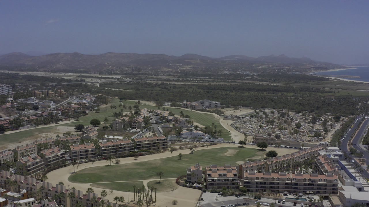 Opening aerial frame over Los Cabos with golf greens, sand traps, red-roofed residences, a road corridor, distant coast, hazy hills, and no visible people.