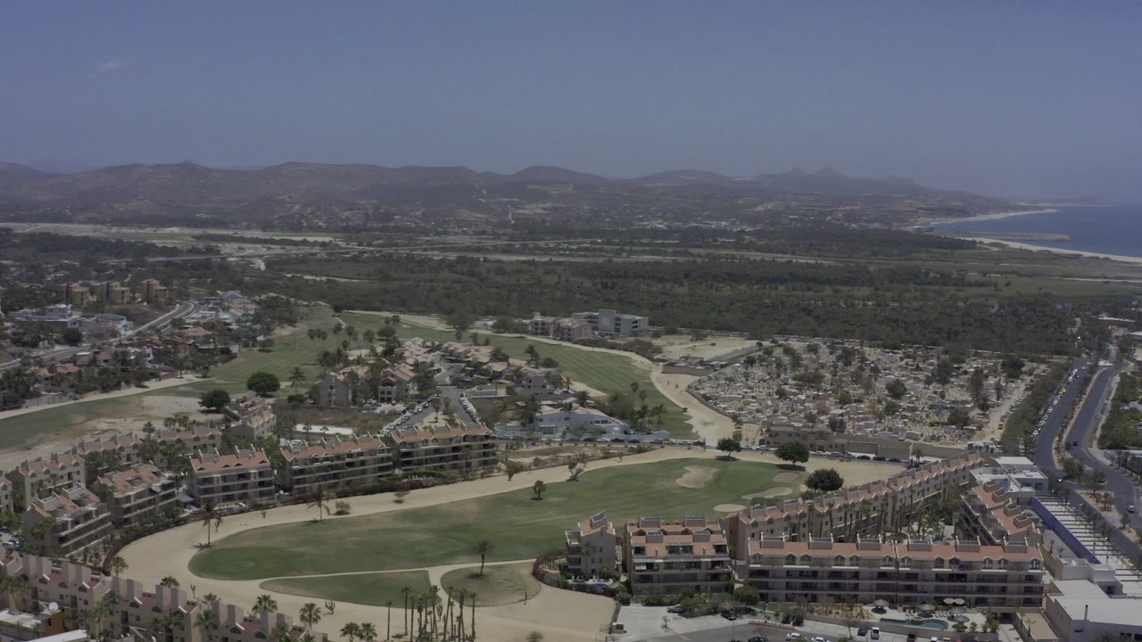 Wide aerial over golf course, resort-style buildings, road, palms, coastline, and hills.