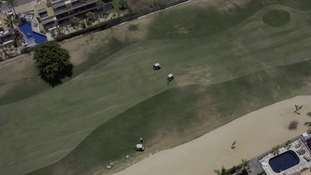 Top-down Los Cabos, Mexico aerial frame showing a green golf fairway, pale sand bunker, two small carts, a large tree, adjacent buildings, blue pools, and no visible people.