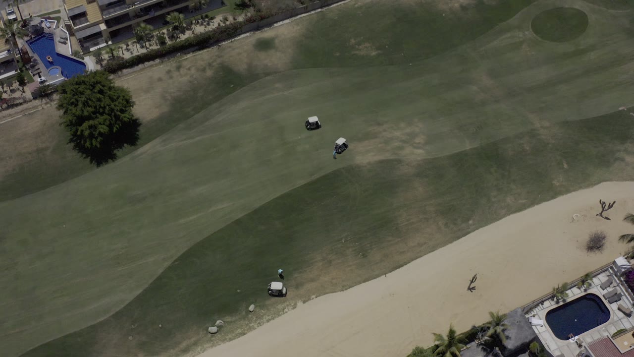 Overhead view of fairway grass, sand bunker, carts, tree, pools, and buildings.