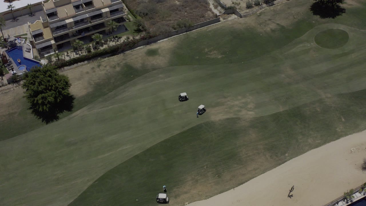 Overhead aerial view of a Los Cabos golf course with fairway grass, two golf carts, a sand bunker, a large tree, edge buildings, and small distant people visible.