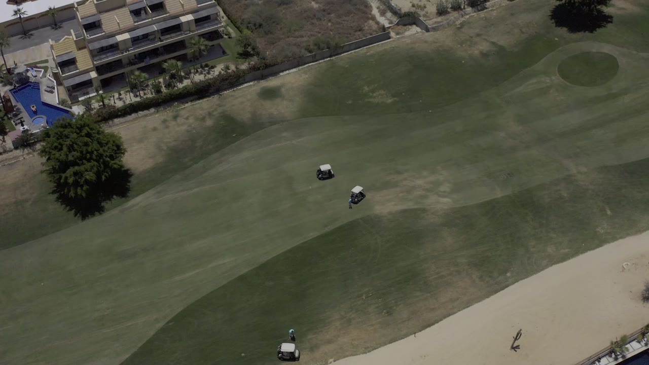 Aerial frame from Los Cabos showing two golf carts on a patchy green fairway, with a sand bunker at lower right and buildings along the upper edge.