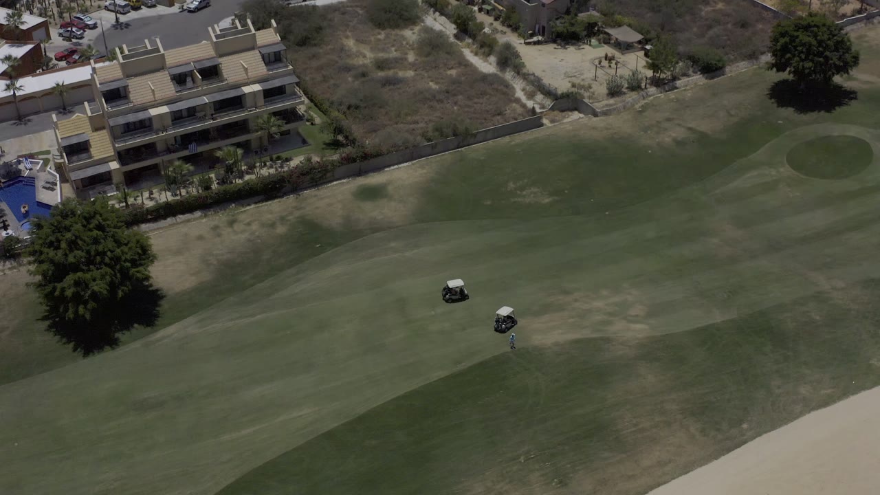 A Los Cabos aerial view holds on the green fairway, tan worn turf, two golf carts, a broad shade tree, condo balconies, and no clearly visible people.