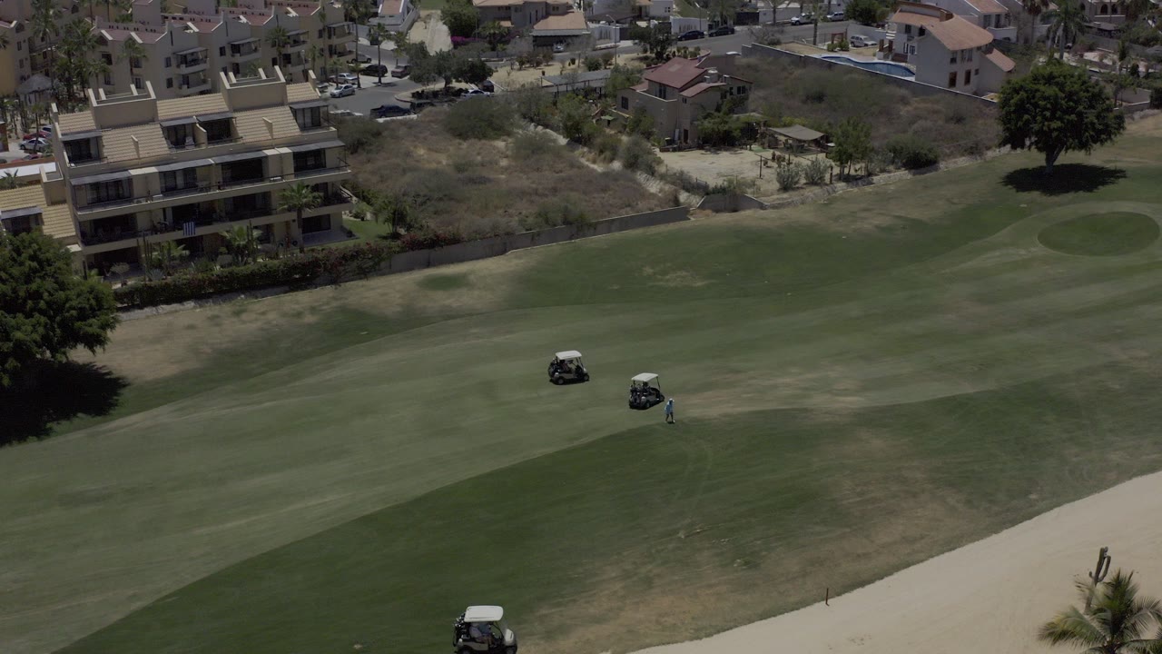 Elevated Los Cabos golf-course view with three carts spread across the fairway, a sand path at the bottom edge, and buildings beyond the rough.