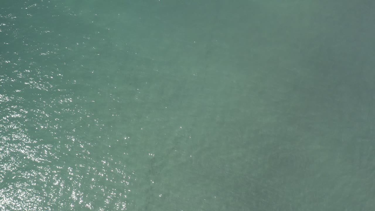 Top-down aerial view of green-blue open sea in Los Cabos, Mexico, with bright glare along the left side and no visible people.