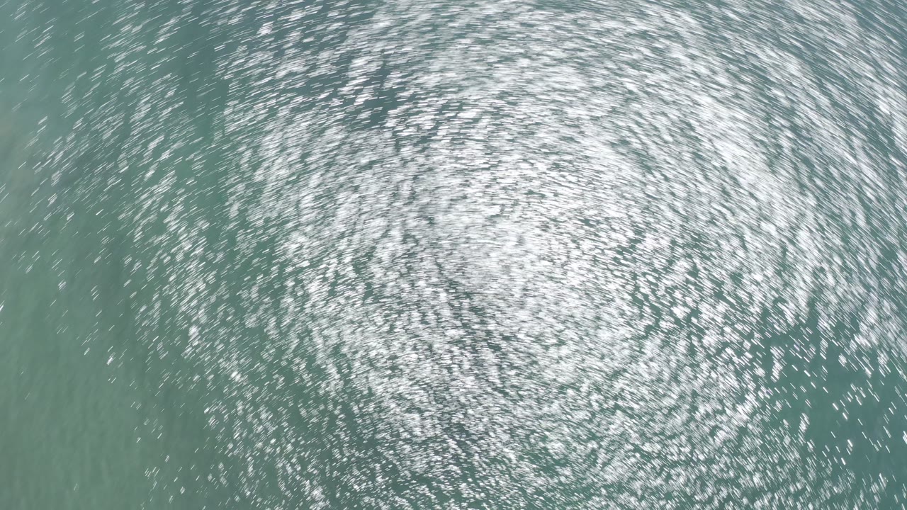 Top-down aerial view of green ocean water in Los Cabos with dense silver sun reflections and no visible people.