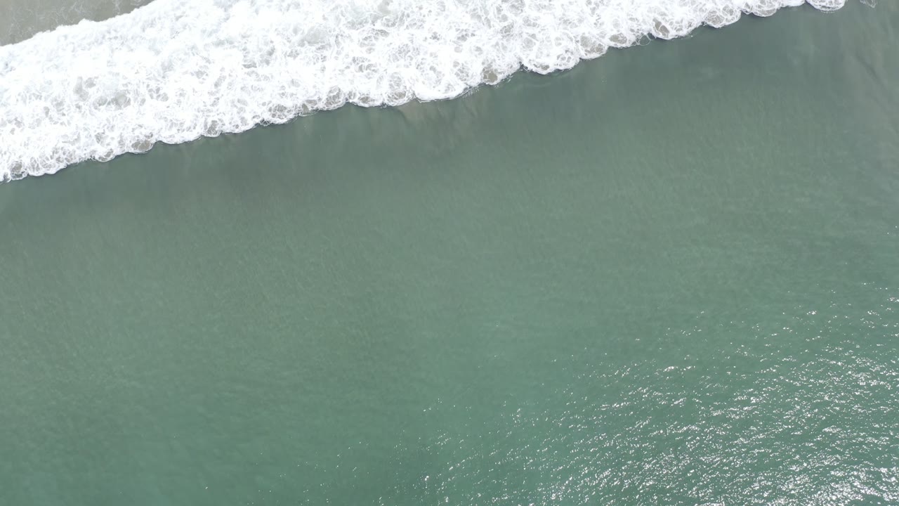 Aerial Los Cabos shoreline frame with white foam stretching diagonally across green water and a thin strip of pale sand near the top, with no visible people.