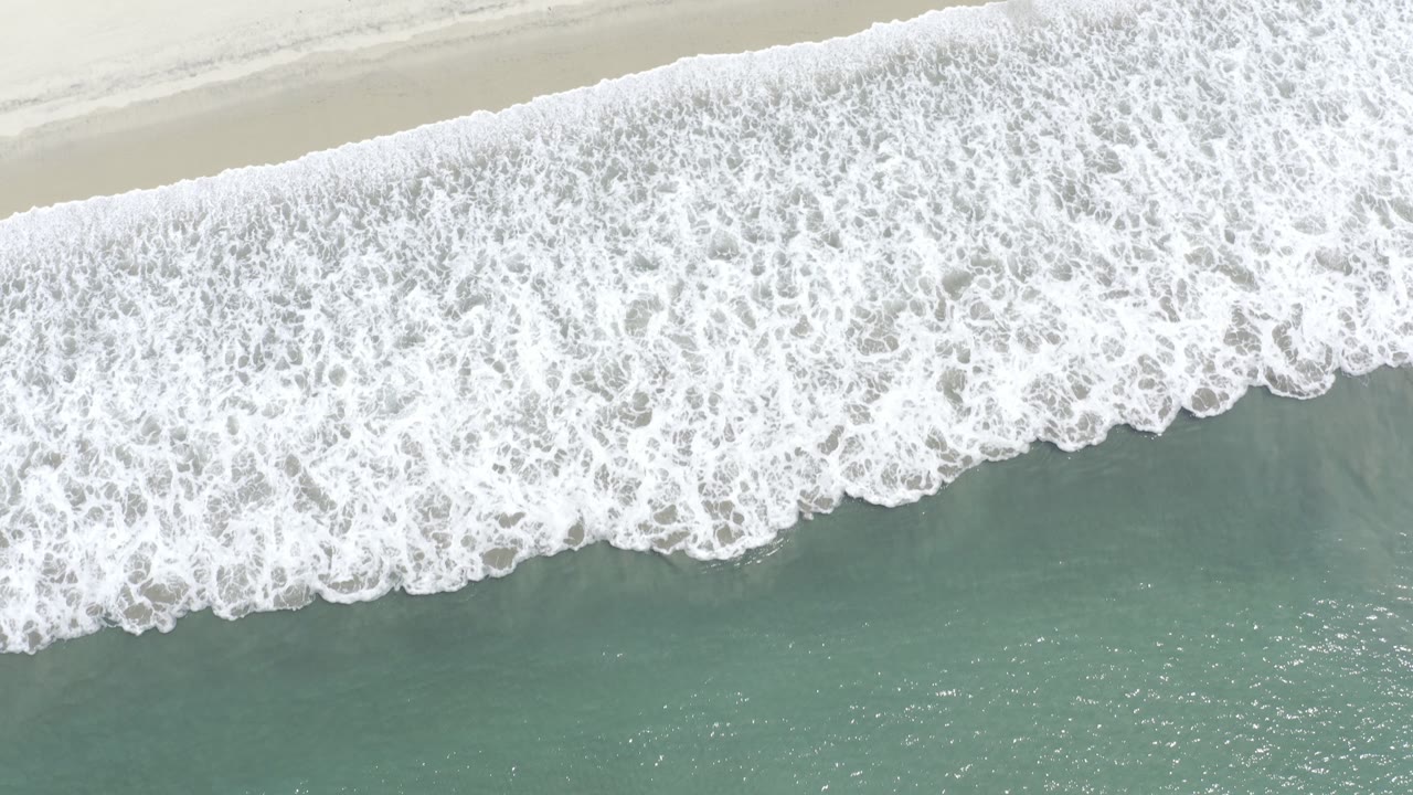 Overhead aerial view in Los Cabos, Mexico, showing a wide band of white surf meeting pale sand above green ocean water, with no visible people.