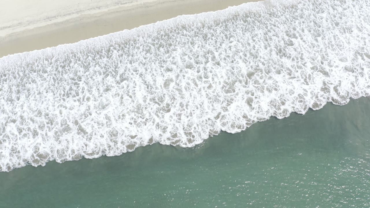 Aerial frame over Los Cabos surf with dense white foam spread diagonally across pale sand and muted green water, with no visible people.