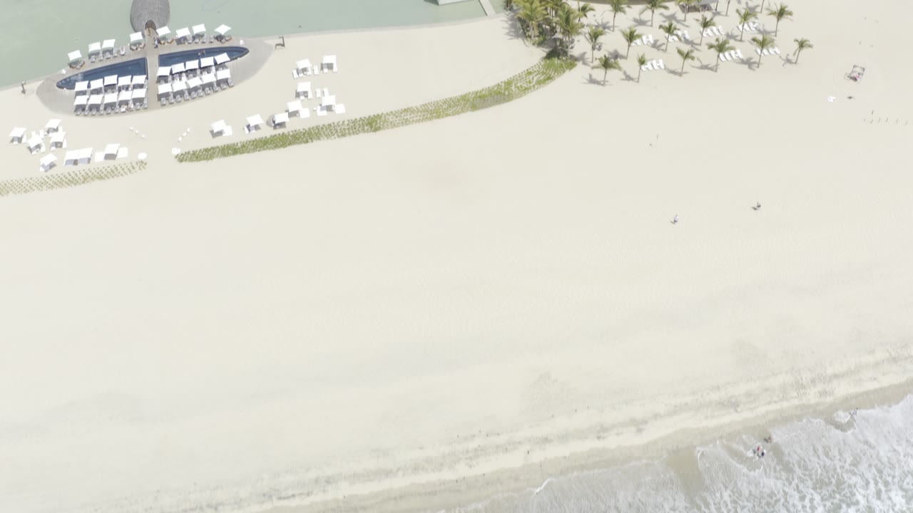 High aerial frame over a Los Cabos beach showing pale sand, shallow turquoise water, a small surf line, and a resort lounge area near the top edge, with no visible people.