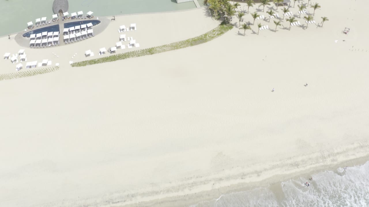 High aerial view of the Los Cabos shoreline with pale sand filling most of the frame, resort chairs near the upper edge, and faint surf washing in from below, with no visible people.