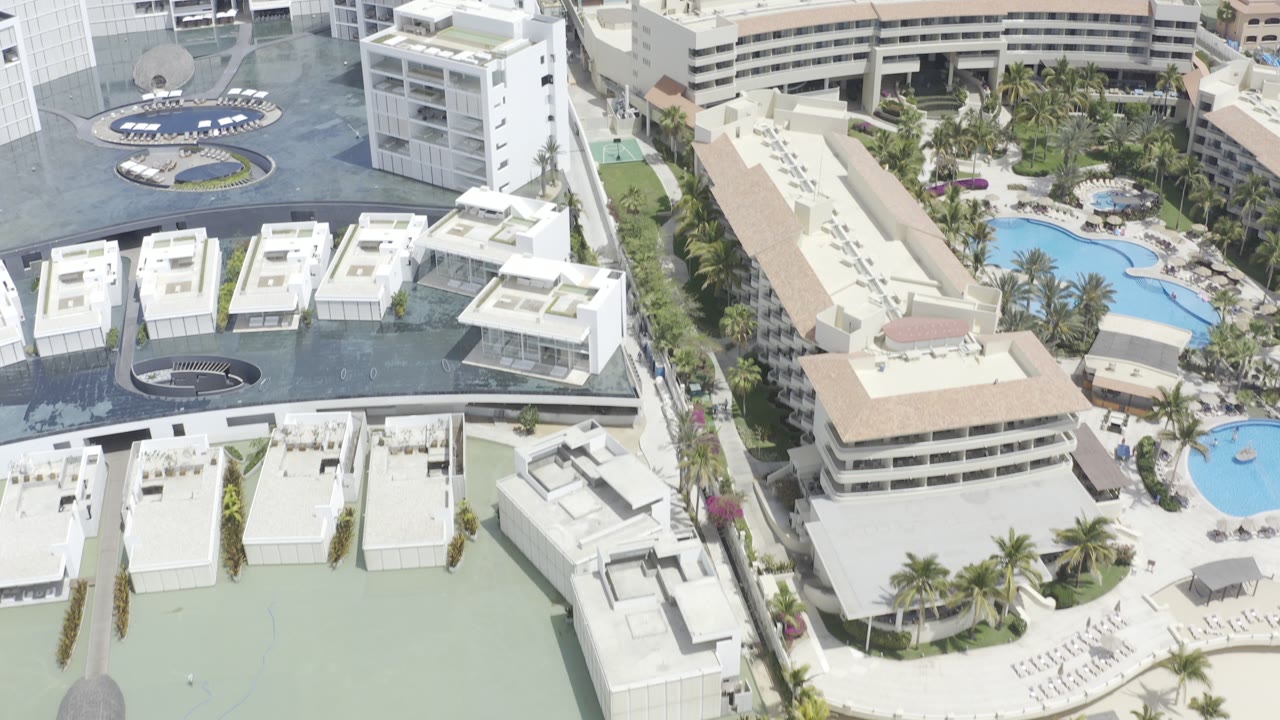 Overhead view of white hotel and condo buildings, blue pools, palm rows, and paved driveways in Los Cabos, Mexico, with no visible people.