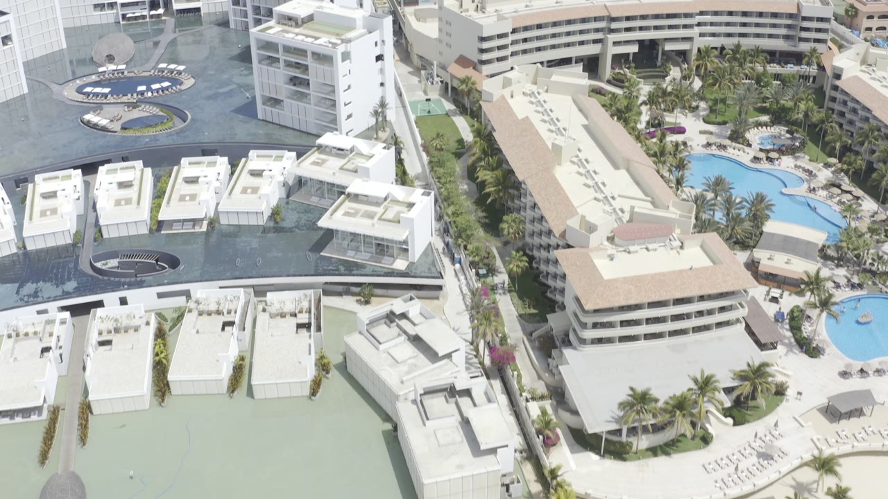 Aerial frame over Los Cabos, Mexico showing resort roofs, white condo blocks, curved blue pool shapes, and palm landscaping, with no visible people.