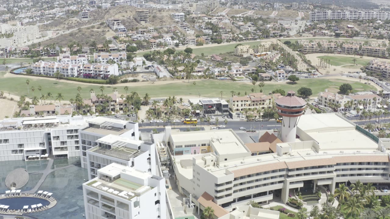 Aerial view over large white buildings, roads, green fairways, hillside housing, and hazy mountains.