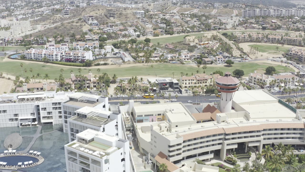 Los Cabos aerial view with resort-scale white buildings low in frame, a broad road with small vehicles, green planned landscaping, dense hillside development, distant mountains, and no visible people.