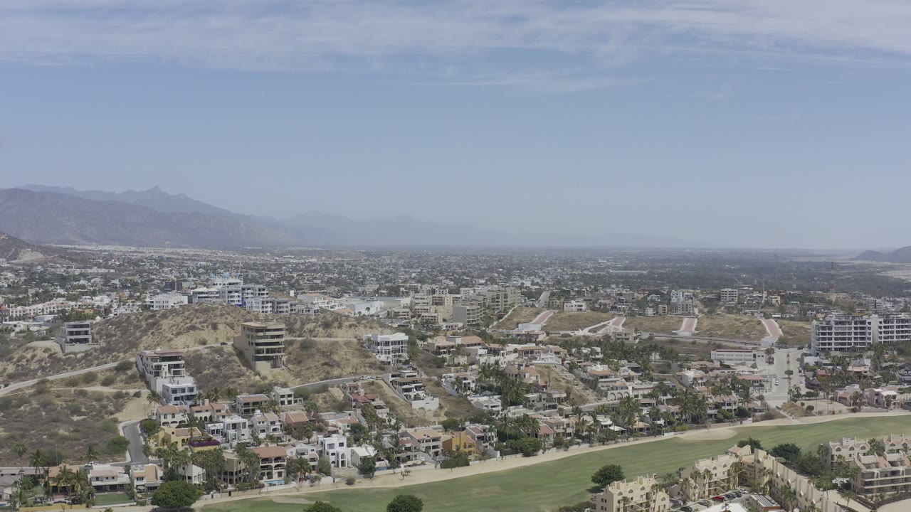 Opening aerial frame of Los Cabos, Mexico, with hillside homes in the foreground, a green strip at the bottom edge, hazy mountains in the distance, and no visible people.
