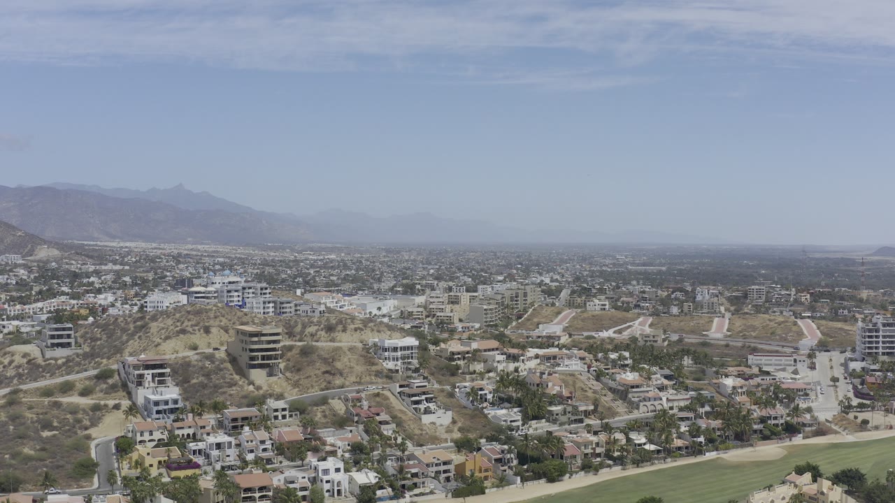 Wide aerial view of hillside homes, dry terrain, distant mountains, and pale sky.