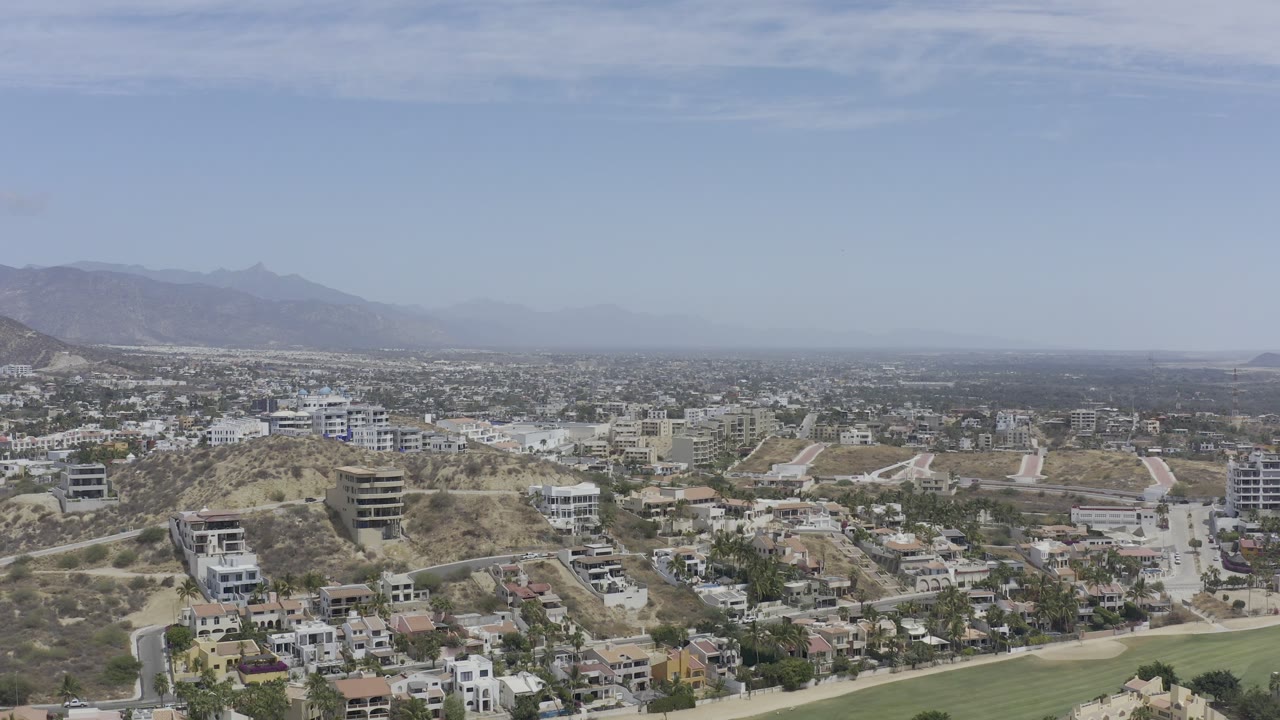 A Los Cabos aerial view shows modern hillside buildings and winding roads in the foreground, a spread of low development in the distance, hazy mountains, and no visible people.