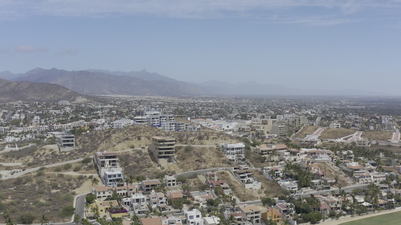 Aerial frame over Los Cabos, Mexico, with hillside homes, winding roads, dry scrub slopes, hazy mountains, pale sky, and no visible people.