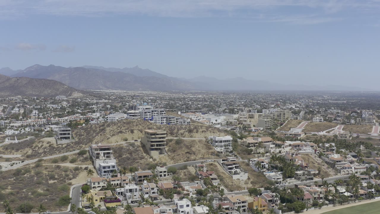 Wide Los Cabos aerial view showing white residential buildings on dry hills, road curves, city blocks beyond, distant mountains, and no visible people.