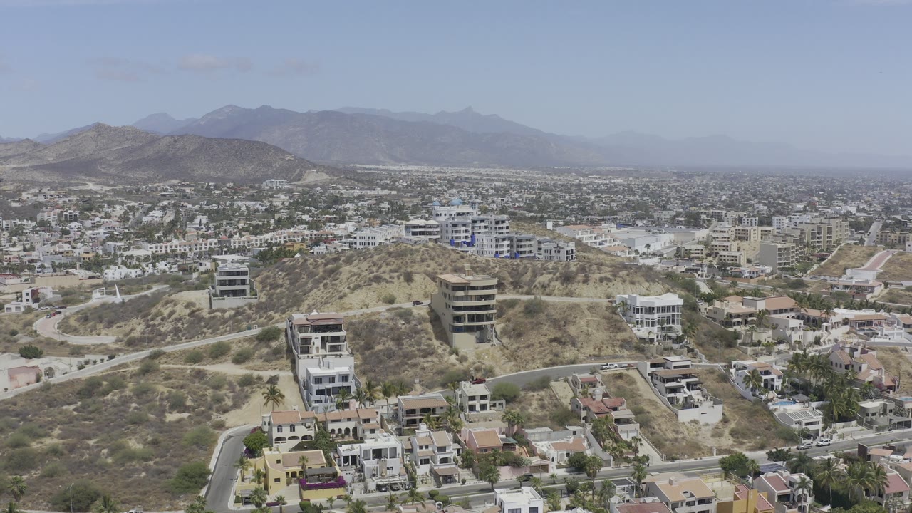 Opening aerial frame over Los Cabos with hillside houses in the foreground, low-rise city buildings beyond, hazy mountain ridges in back, and no visible people.