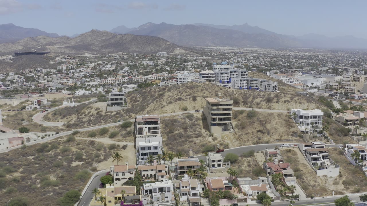 Los Cabos aerial view with white villas, tan scrub-covered slopes, roads curving through the hills, hazy mountains, and no visible people.