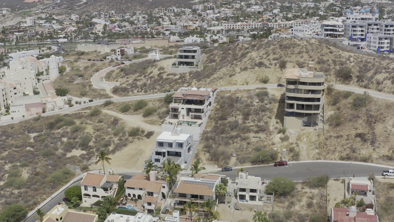 Aerial view in Los Cabos, Mexico with a round-front hillside building, nearby modern homes, a pale dirt track, a curved paved road, and no visible people.