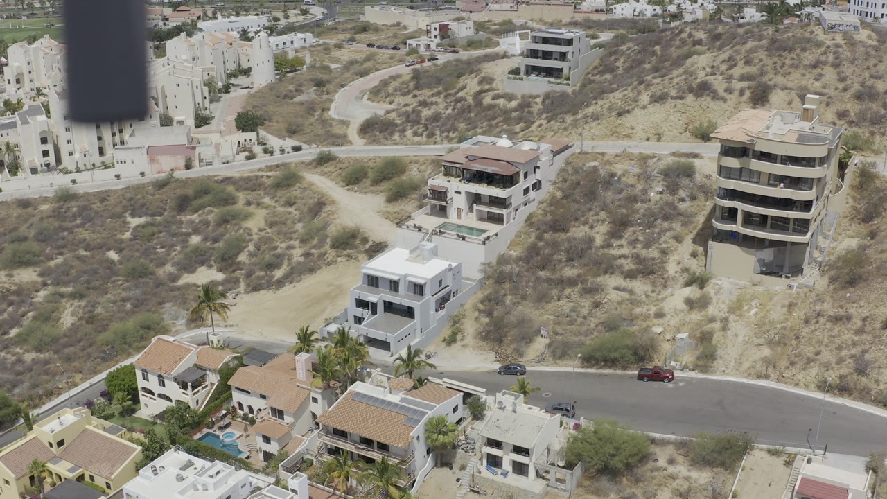 Drone frame over Los Cabos showing white homes, tan-roofed houses, hillside dirt paths, a curved paved street, small pools, and no visible people.
