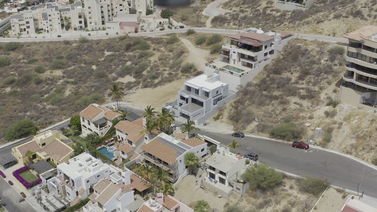 Aerial view over Los Cabos homes, a curved paved road, dry hillside lots, and modern buildings in bright sun with no visible people.