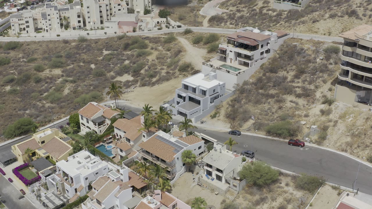 High aerial frame of Los Cabos residential roofs, palm trees, a road bend, parked vehicles, and pale hillside terrain with no visible people.