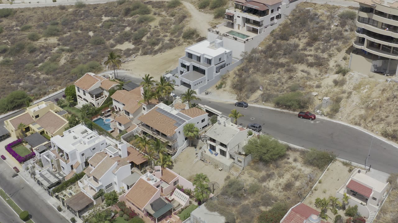 Aerial view in Los Cabos showing white and tan residential buildings, palm trees, a curved paved road with vehicles, dry brush, and no visible people.