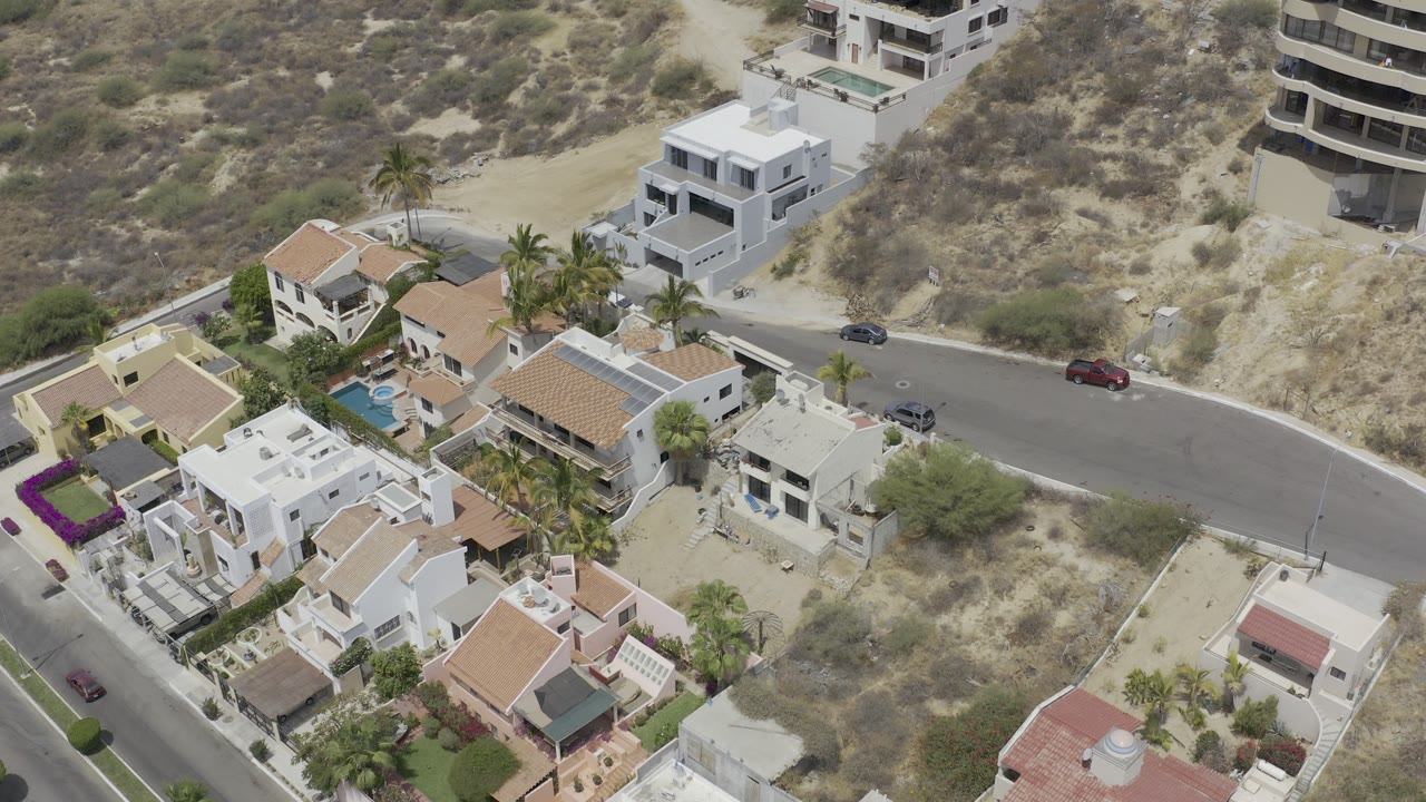 Aerial view over Los Cabos, Mexico rental houses with terracotta roofs, a curved road, dry hillside lots, palm trees, parked cars, and no visible people.
